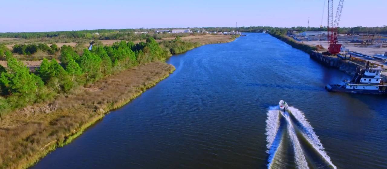 Aerial view of a small boat cutting a white wake up a wide canal, marsh and pines on one bank and a waterfront with a red crane and a tug on the other