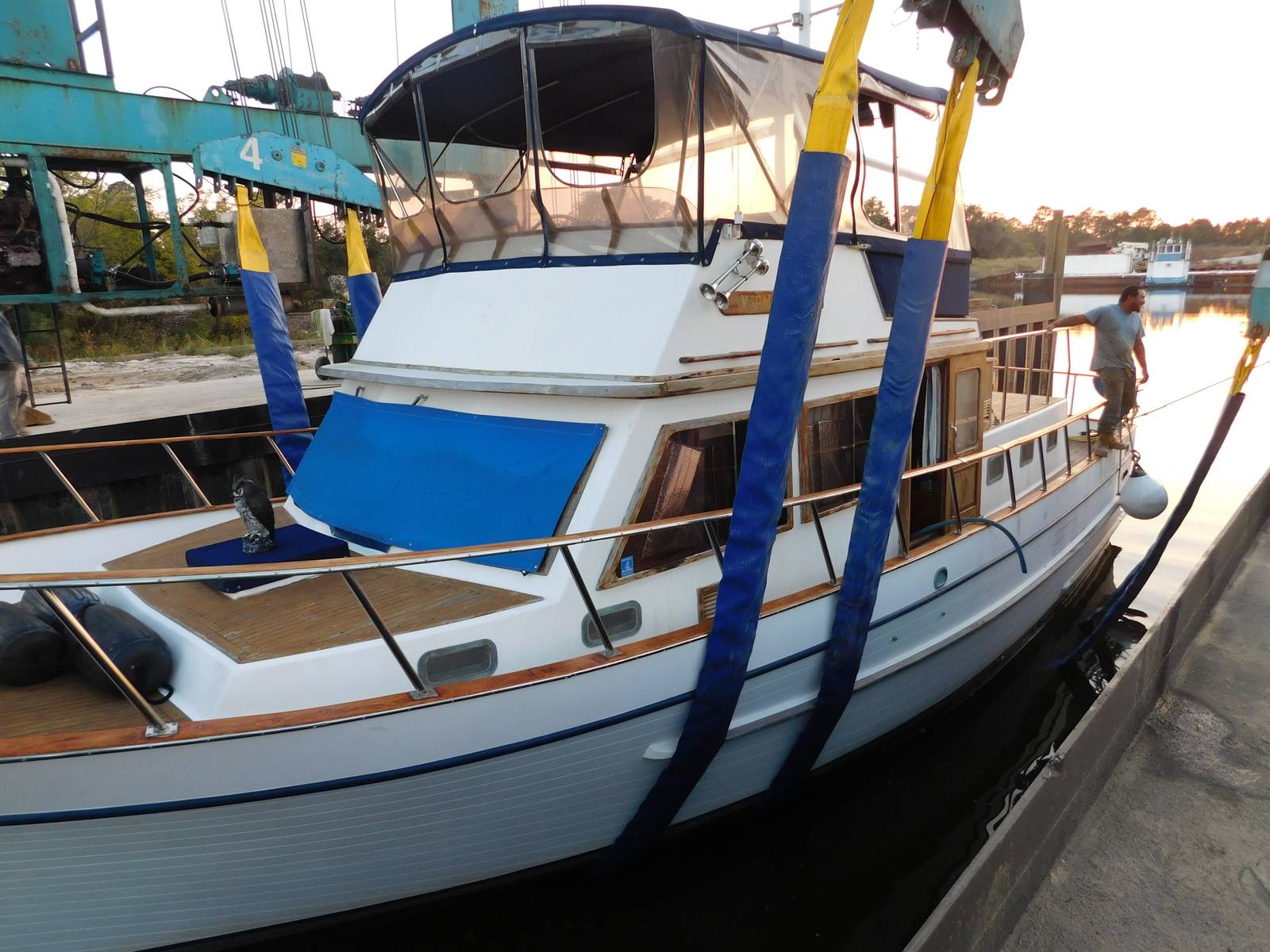 White trawler-style motor yacht with teak trim and blue canvas held in the blue and yellow slings of a teal travel lift at the haul-out slip, a worker on the bow, canal and tree line at sunset behind